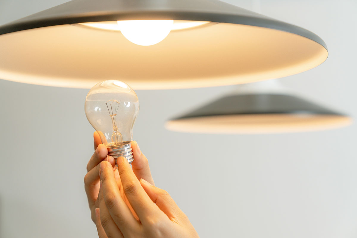 A close-up shot of a person's hands holding a new lightbulb up to a farmhouse-style pendant light that is turned on and features a different style of lightbulb.