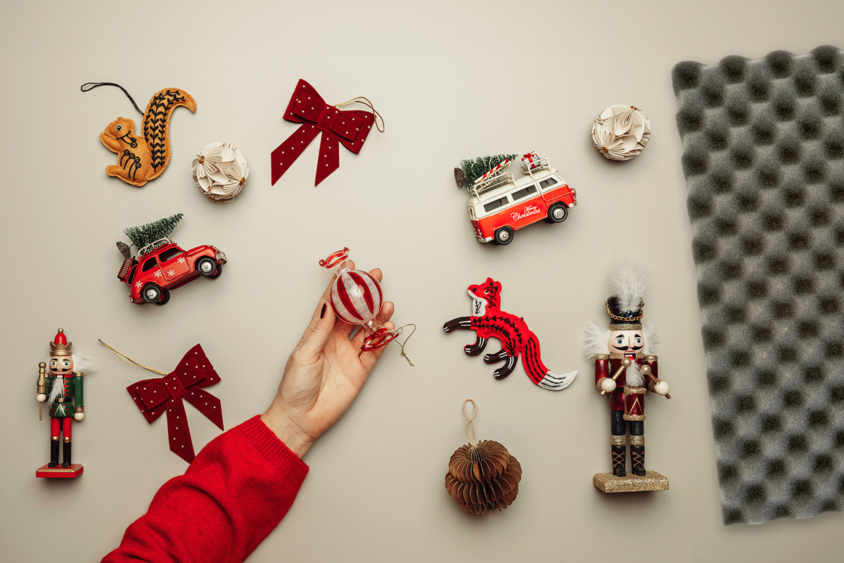 Anonymous person wearing a red sweater holding a peppermint candy-shaped glass ornament in their hand. On the same surface are other delicate-looking Christmas ornaments and a piece of gray egg crate foam.