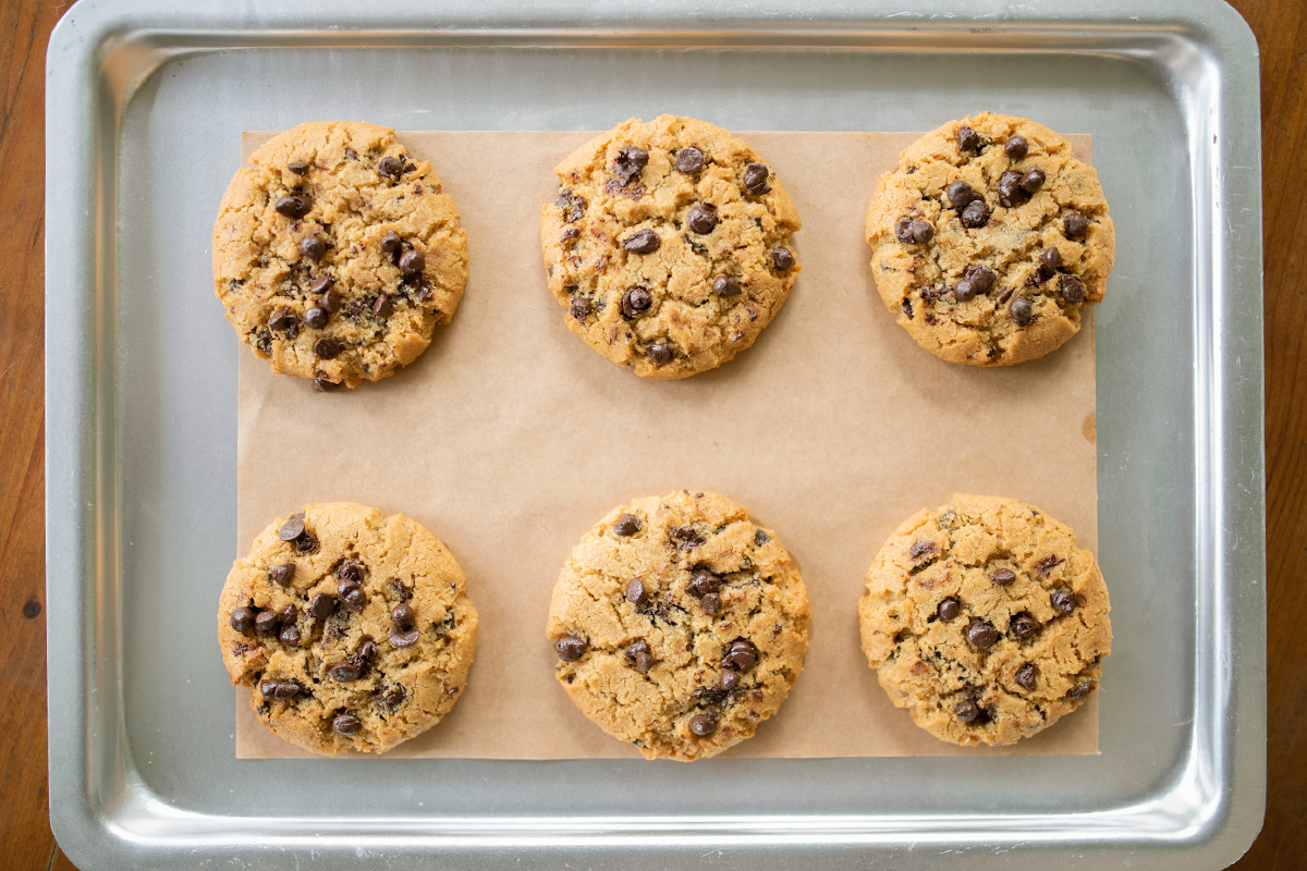 An overhead shot of a half-sheet baking pan with six perfectly round chocolate chip cookies on a piece of parchment paper.