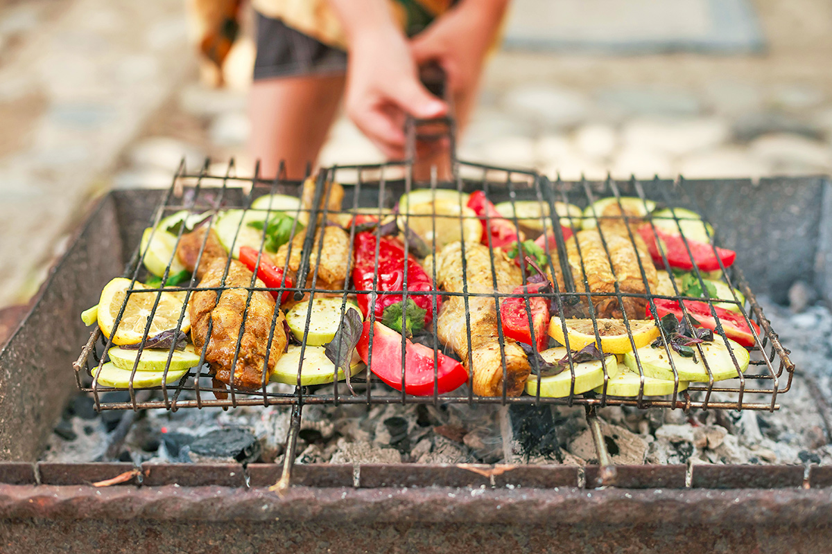 Person using a wire grilling basket with a handle so it can be flipped on a charcoal grill. Inside the basket are veggies, chicken drumsticks, and lemon slices.