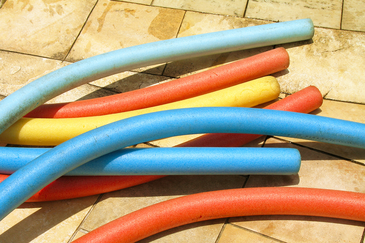 Seven pool noodles of various colors on a wet outdoor patio with tile flooring.