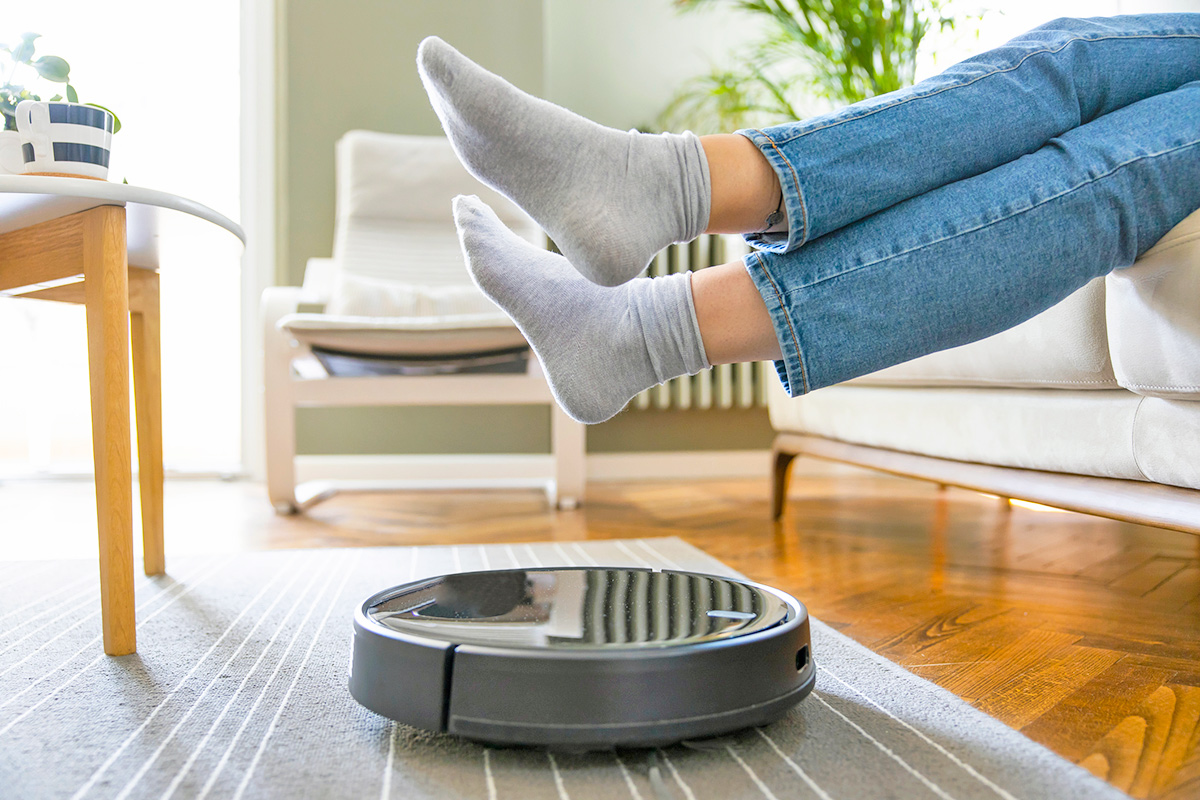 Anonymous person sitting on a couch with their feet up so a robot vacuum cleaner can pass by.