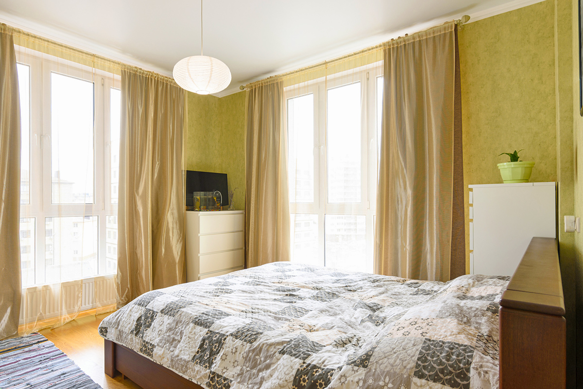 A chartreuse-painted bedroom with sheer gold curtains covering both large windows. The bed in the middle of the room has a quilt on it.