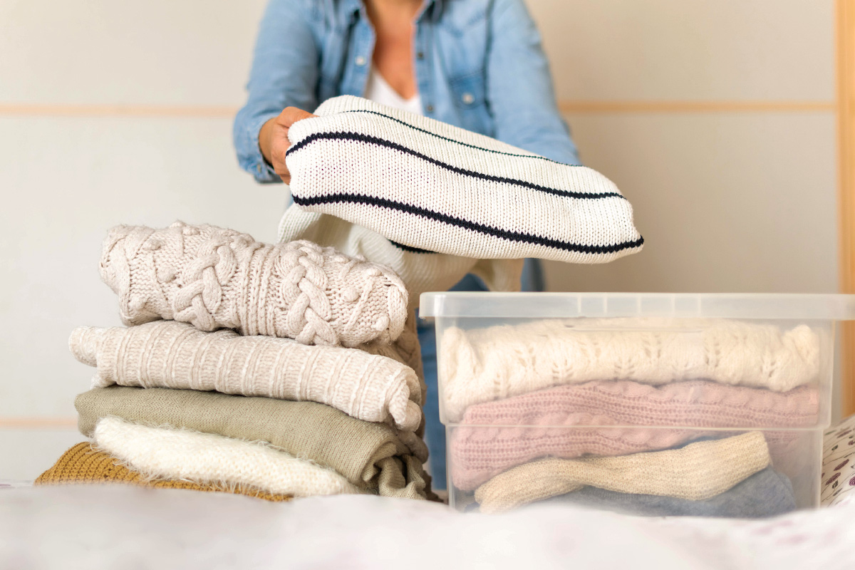 Anonymous woman putting folded sweaters into a clear storage container.