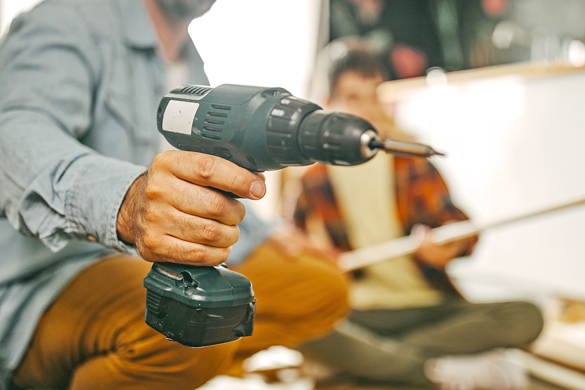 A father holding up an electric drill to the camera with his son in the background, out of focus.