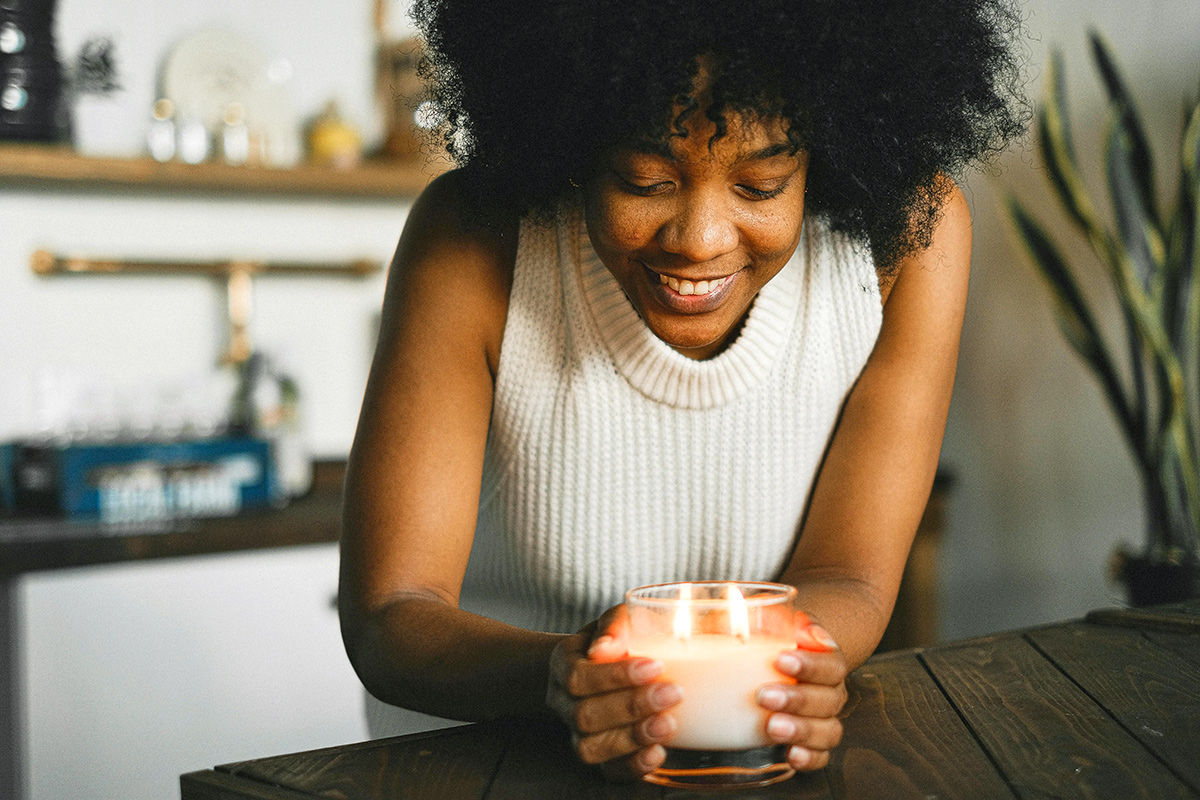 A young woman wearing a white knit tank top leaning over a lit, double-wick candle in a glass jar.