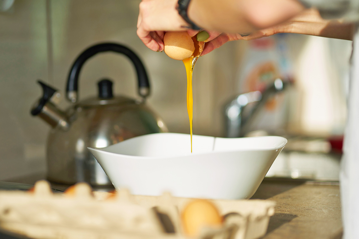 Anonymous person cracking an egg into a bowl. Next to the bowl is an open carton with a few remaining eggs.
