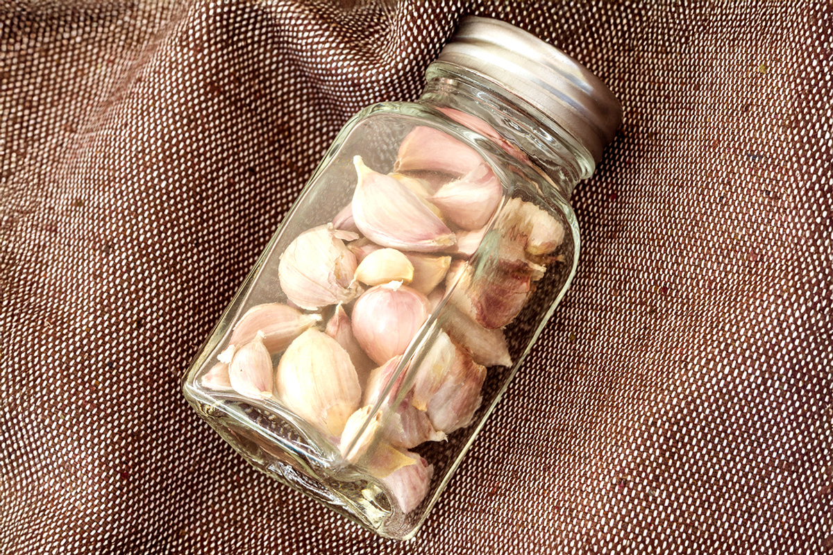 Unpeeled cloves of garlic in a glass jar. The jar is on its side, laying on brown cloth.