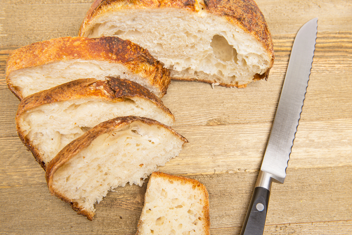 A loaf of crusty bread on a wooden cutting board that's been sliced with a serrated knife.