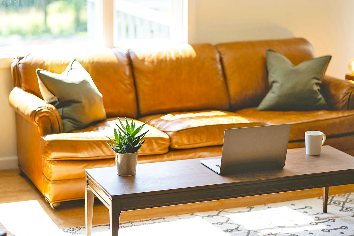 Tan leather couch with a long walnut coffee table in front of it. On the table is a laptop, plant, and coffee cup.