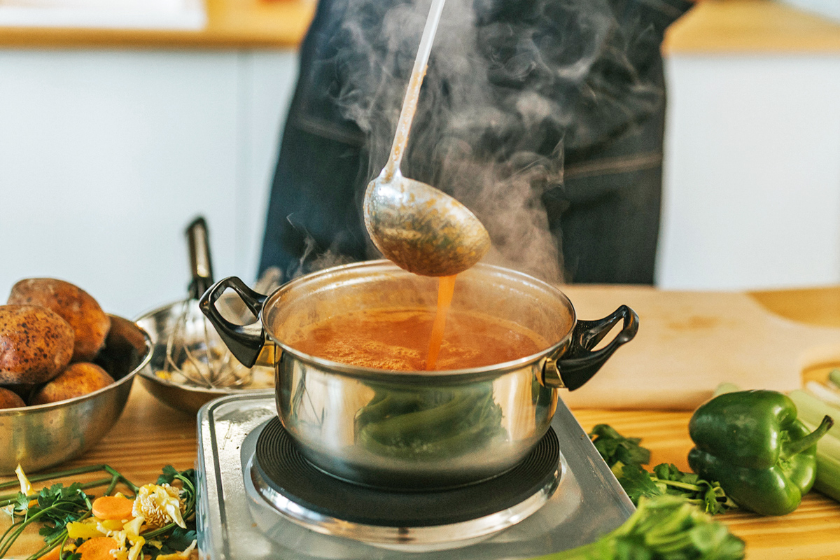 Anonymous person cooking a pot of thin-looking soup on a hot plate. They're using a ladle to mix it.
