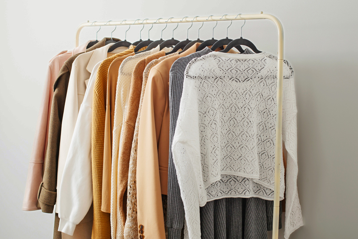 A freestanding clothing rack in a gold finish with women's clothing on it, mostly sweaters and coats.