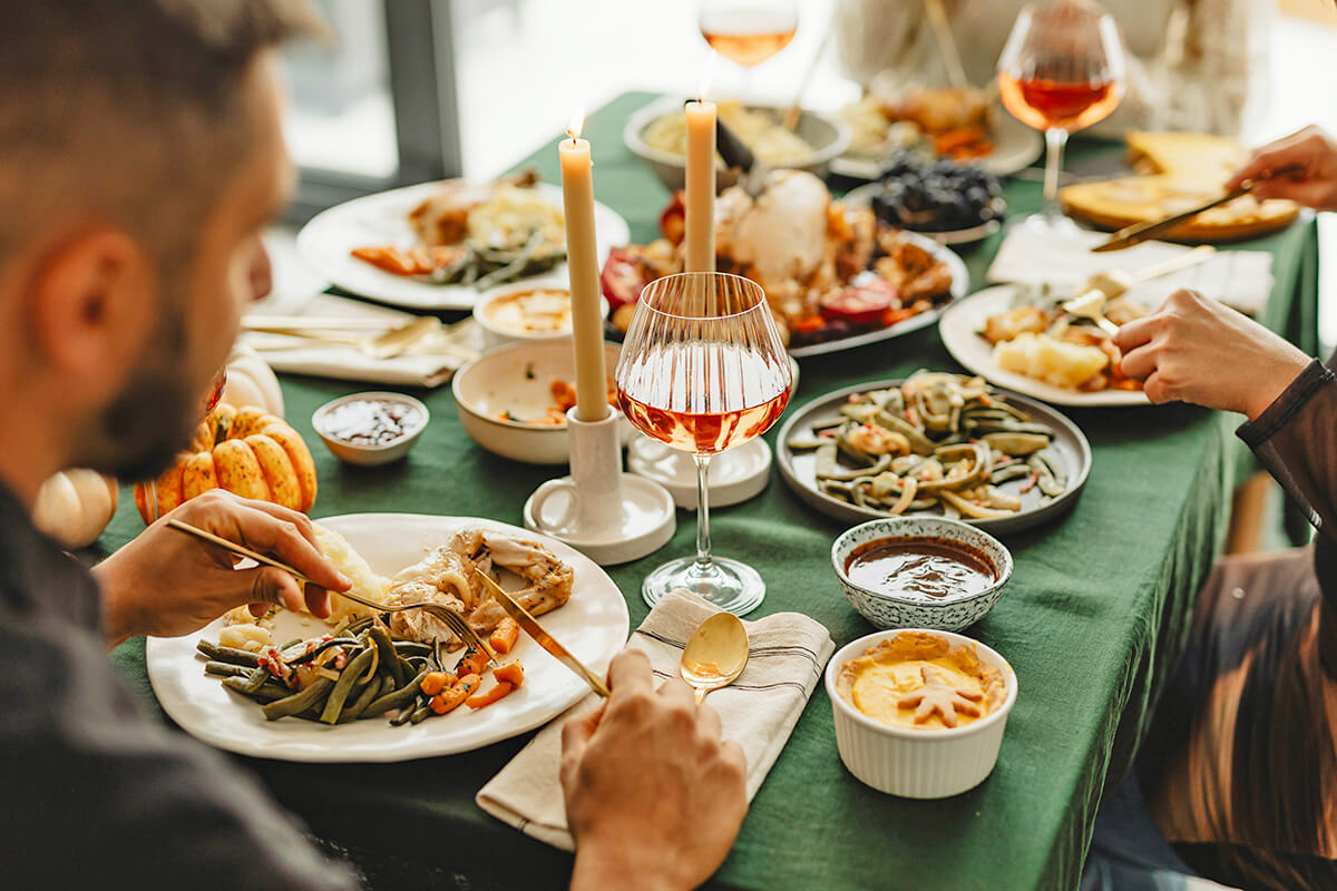 Three people sitting at a dining table with a green tablecloth on it. They are eating Thanksgiving foods such as turkey, mashed potatoes, carrots, and green beans.