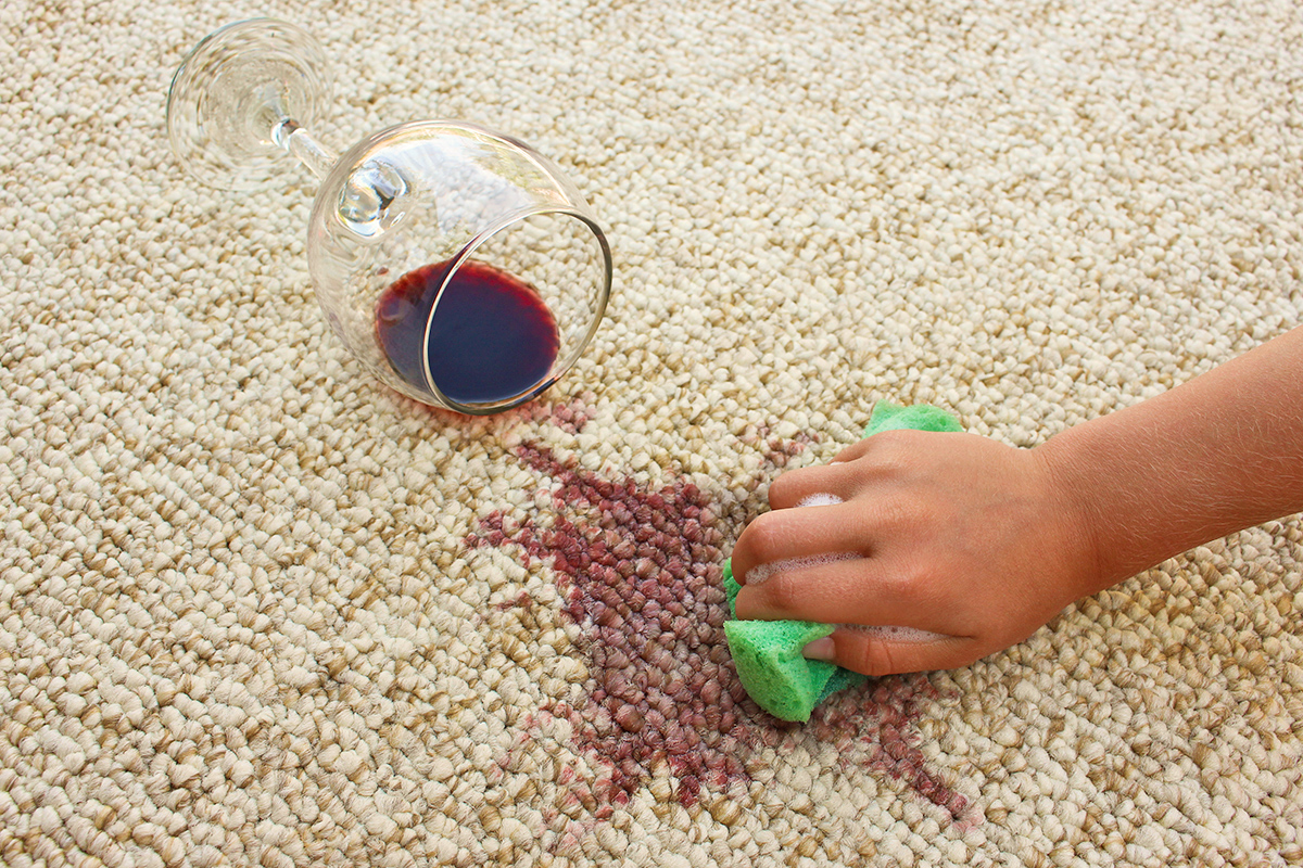 A spilled glass of red wine on beige-colored carpet. An anonymous person is attempting to clean it with a soapy sponge.